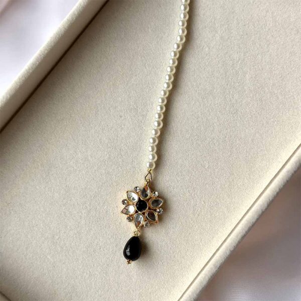 Kundan Maang Tikka with intricate gold plating, floral embellishments, white stones, and pearls, displayed on a white textured surface with green leaves and red fabric