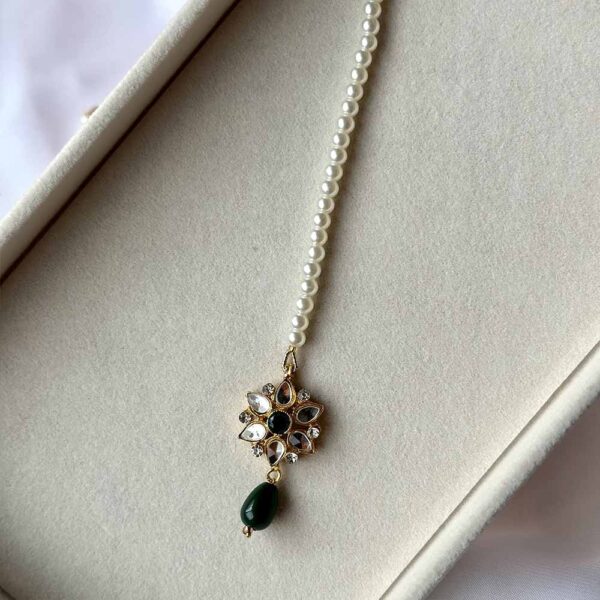 Kundan Maang Tikka with intricate gold plating, floral embellishments, white stones, and pearls, displayed on a white textured surface with green leaves and red fabric