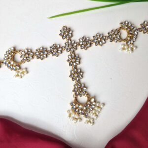 Traditional Kundan Matha Patti with intricate gold plating, white stones, and pearls, displayed on a textured surface with red fabric and green leaves.